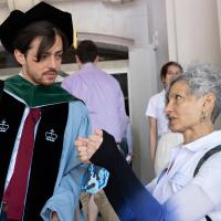 A doctoral graduate in their full regalia is looking at a woman in a white polo shirt as she is speaking